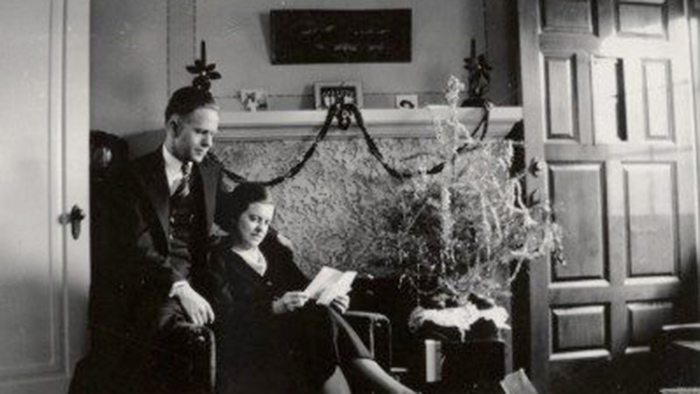 man sitting on armrest of chair with woman reading n front of fireplace decorated for Christmas