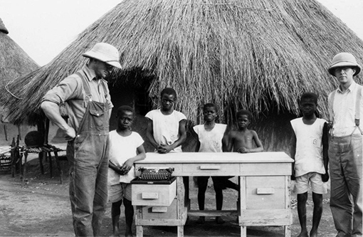 photo of two men and five Camaroon children by desk in front of thatched roof hut