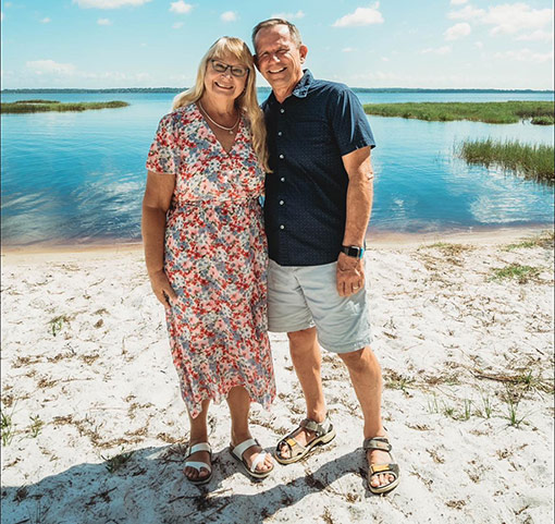 woman in print dress with man in shorts and polo shirt standing on beach with lake in background