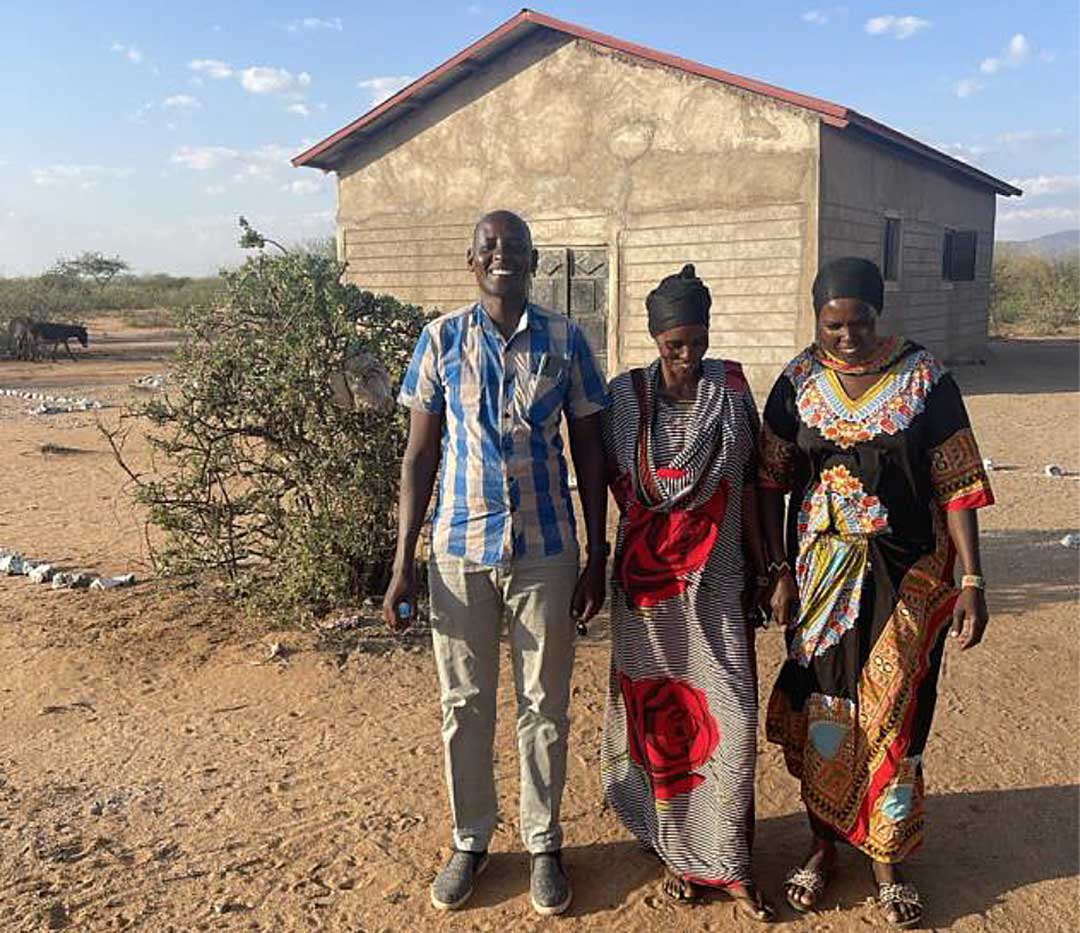 man and two women standing in dirt yard in front of primitive house in Kenya