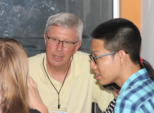 man in yellow shirt conversing with woman and man in classroom with blackboard in background