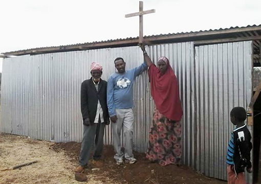 two men and one woman and child standing in front of corrugated steel exterior wall of church in Kenya, pointing up to cross on roof