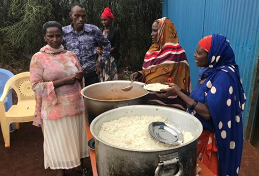 three women and one man dressed in colorful African clothing dishing up food from huge kettles of rice and gravy