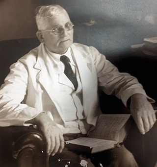 older man wearing white doctor's coat, seated in chair with Bible open on his lap