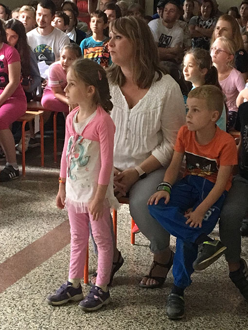 girl wearing pink shirt and pants with mother and brother sitting behind her at group function