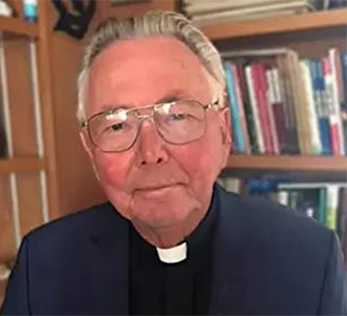 older man wearing black suit and shirt with clerical collar sitting in front of bookshelf