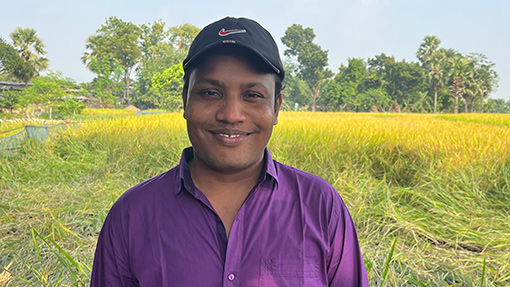 man wearing purple shirt standing in field of grain with trees in background
