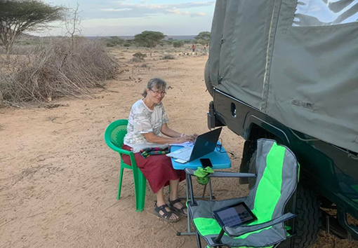 Woman sitting on green lawn chair working at laptop computer on blue TV tray beside lorry in desert region of Kenya