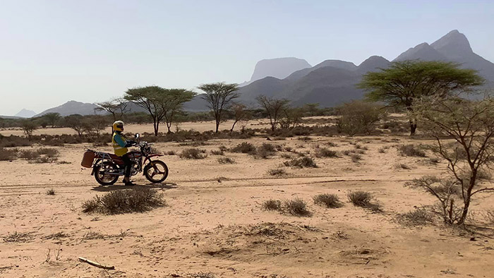 man riding motorcycle in desert region of Kenya with baobab trees and mountains in background