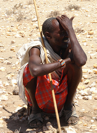 elderly Kenyan man wearily squatting on rocky desert ground with elbows on knees and palm on forehead