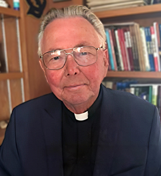 photo of man wearing navy suit coat and white clerical collar sitting in front of bookcase