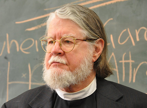 profile view of man with gray hair, beard, mustache, wearing black robe with clerical collar