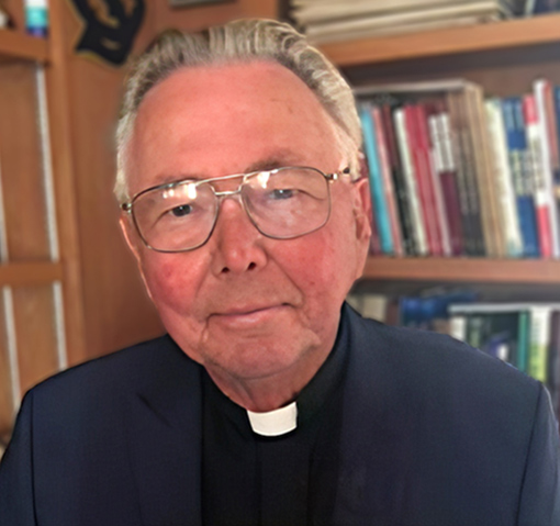 man with gray hair and glasses wearing clerical collar with bookcase in background
