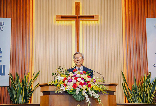 man standing behind flowers on pulpit with large cross in background