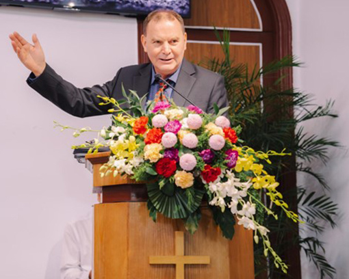 man in suit with right arm extended standing behind flowers on pulpit in a church