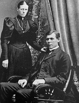 vintage photo of woman in dark dress standing beside seated husband