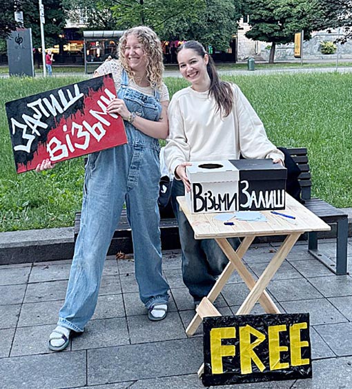 woman wearing overalls displaying banner standing next to woman in whit sweatshirt standing behind display table